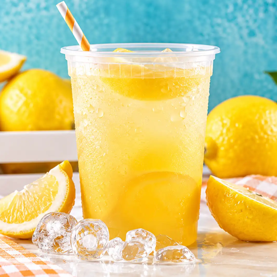 Chilled lemonade in a clear plastic cup with a striped straw, ice cubes, and lemon wedges on a sunny table.