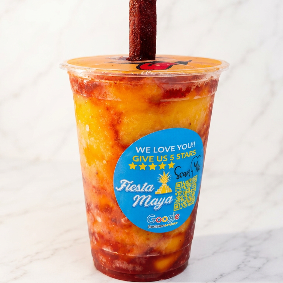 Colorful mango drink with red chamoy swirls, tajín-coated straw, and Fiesta Maya promotional sticker on a marble surface.